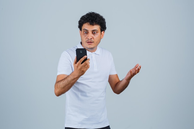 Молодой человек смотрит на мобильный телефон в белой футболке и выглядит озадаченным, вид спереди.