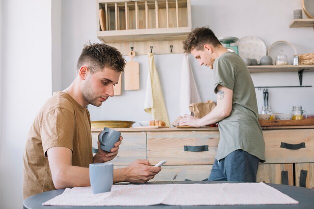 Young man looking at smartphone holding cup while his friend working in kitchen