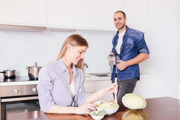 Молодой человек, глядя на улыбающегося жена готовит салат на кухне