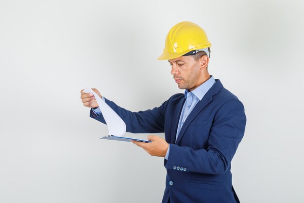 Young man looking through clipboard in suit, safety helmet and looking busy 