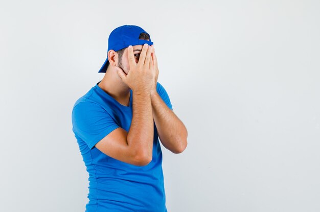 Young man looking through fingers in blue t-shirt and cap and looking frightened