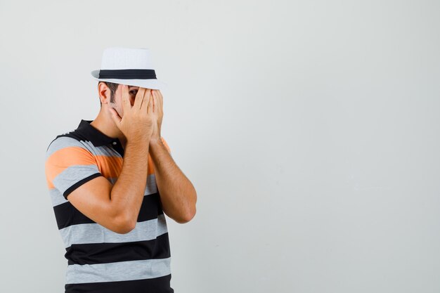 Free photo young man looking through fingers in striped t-shirt,hat and looking scared.  space for text