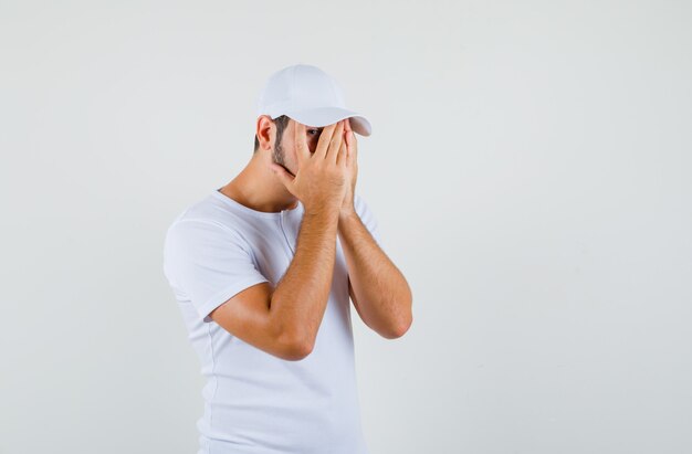 Young man looking through fingers in t-shirt,cap and looking hidden. front view.