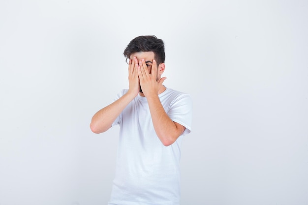 Young man looking through fingers in white t-shirt and looking scared