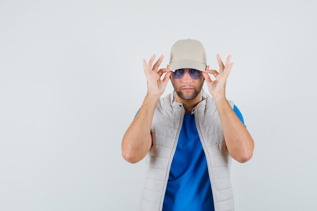 Young man looking through glasses in t-shirt, jacket, cap , front view.