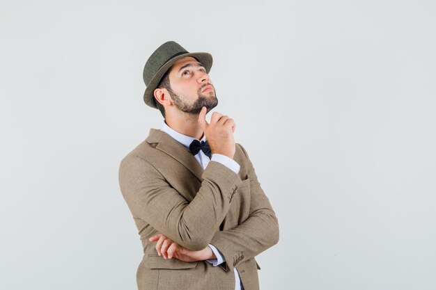 Young man looking up while propping chin on finger in suit, hat and looking thoughtful. front view.