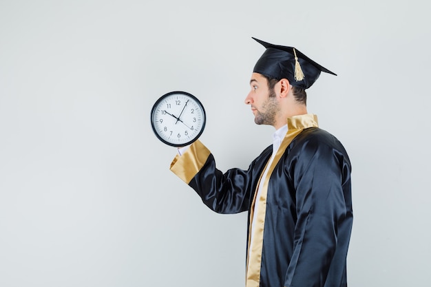 Young man looking at wall clock in graduate uniform and looking focused. .