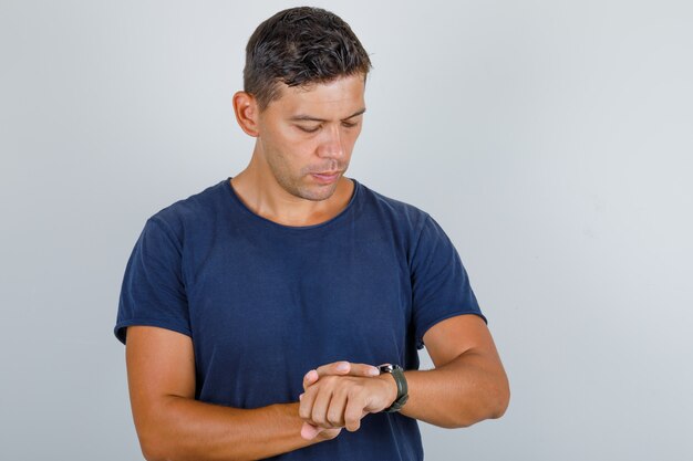 Young man looking at watch on wrist in dark blue t-shirt and looking punctual, front view.