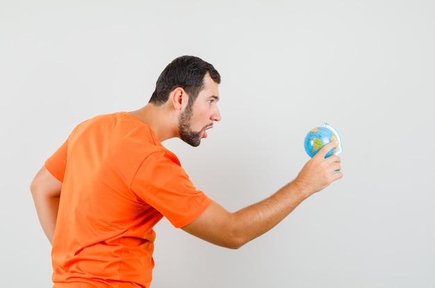 Young man looking at world globe in orange t-shirt and looking focused .