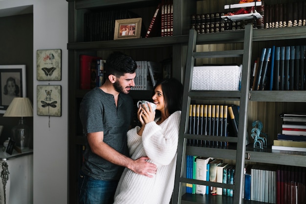 Young man loving her girlfriend holding coffee mug standing in front of bookshelf