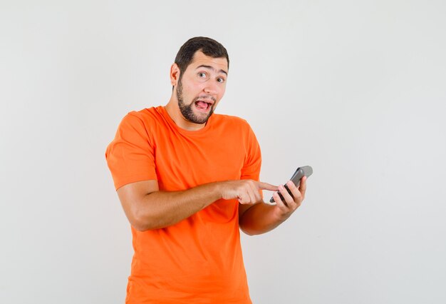 Young man making calculations on calculator in orange t-shirt front view.