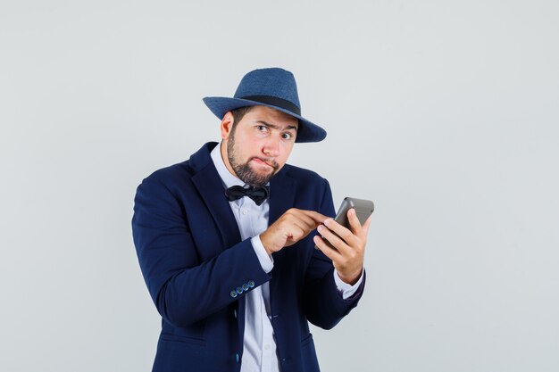 Young man making calculations on calculator in suit, hat and looking cunning. front view.