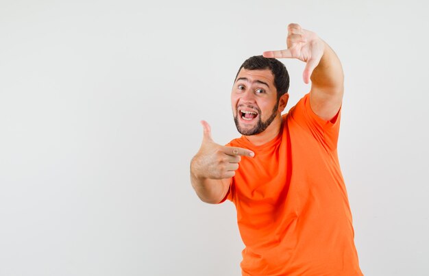 Young man making frame gesture in orange t-shirt and looking cheerful. front view.