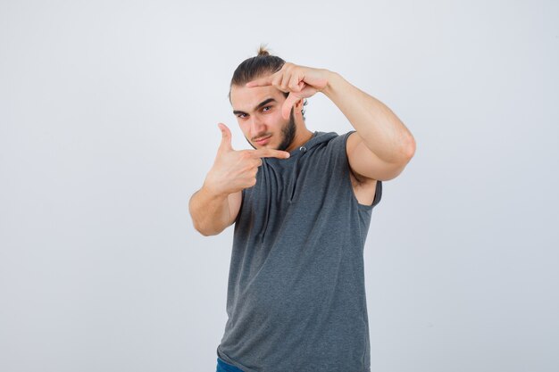 Young man making frame gesture in sleeveless hoodie and looking serious , front view.