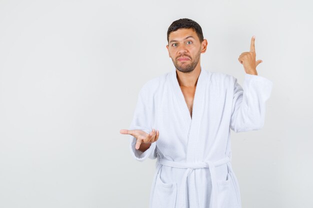 Young man making gun gesture in white bathrobe and looking puzzled , front view.