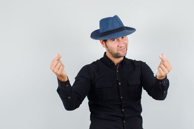 Young man making korean love sign in black shirt, hat and looking merry