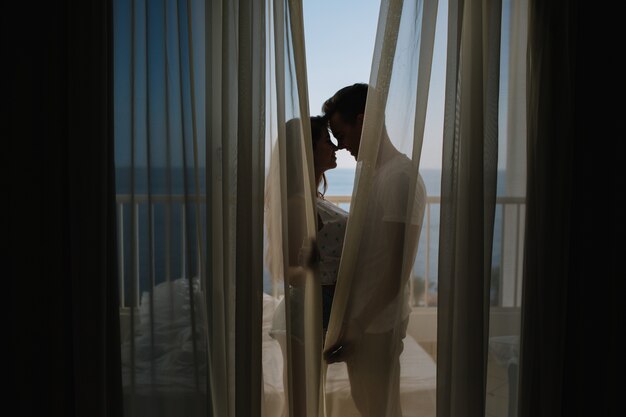 Young man making love confession to his charming girlfriend standing on balcony behind the curtains in morning. Portrait of beautiful couple kissing on terrace after long busy day