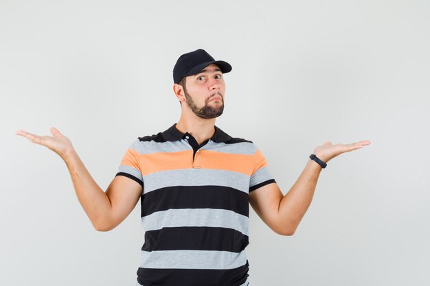 Young man making scales gesture in t-shirt, cap and looking confused. front view.