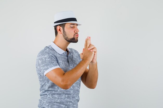 Молодой человек делает жест стрельбы из пистолета в полосатой футболке, шляпе и выглядит сосредоточенным. передний план.