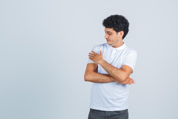 Young man making superiority gesture by wiping shoulder in white t-shirt, pants and looking elegant , front view.