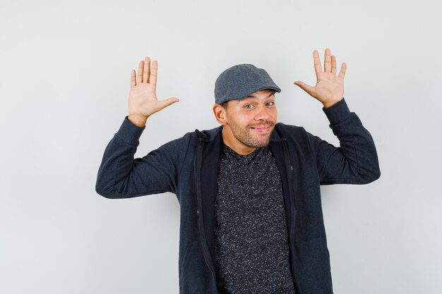 Young man making surrender gesture and smiling in t-shirt, jacket, cap front view.