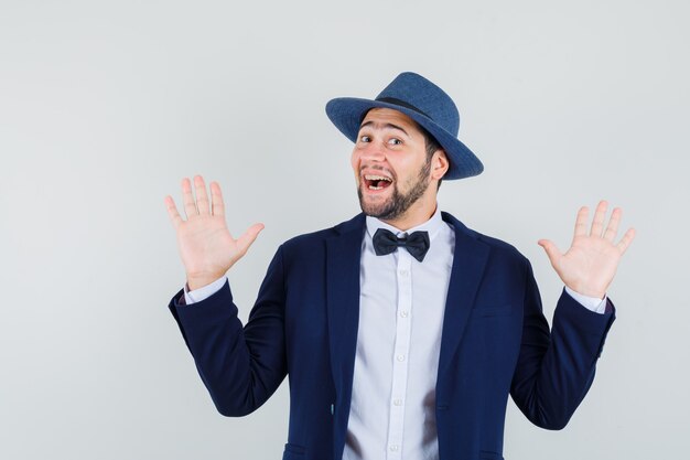 Young man making surrender gesture in suit, hat and looking cheerful. front view.