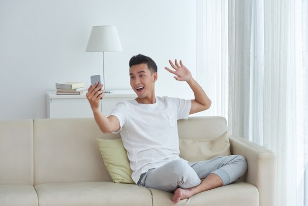 Young man making a video call sitting on the sofa in his living room and giving a greeting wave gesture