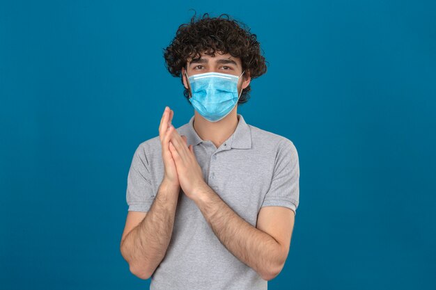 Young man in medical protective mask looking at camera rubbing hands over isolated blue background