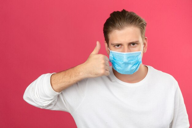 Young man in medical protective mask looking at camera showing thumbs up standing over isolated pink background