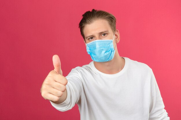 Young man in medical protective mask looking at camera showing thumbs up standing over isolated pink background