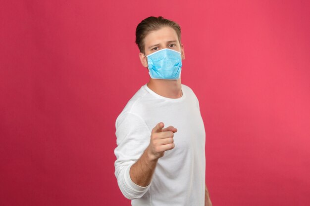 Молодой человек в медицинской защитной маске выглядит уверенно, указывая пальцем на камеру на изолированном розовом фоне