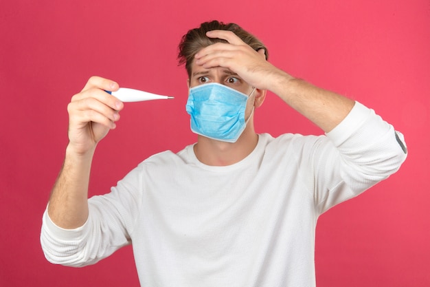 Young man in medical protective mask looking at digital thermometer in panic shocked and surprised over isolated pink background