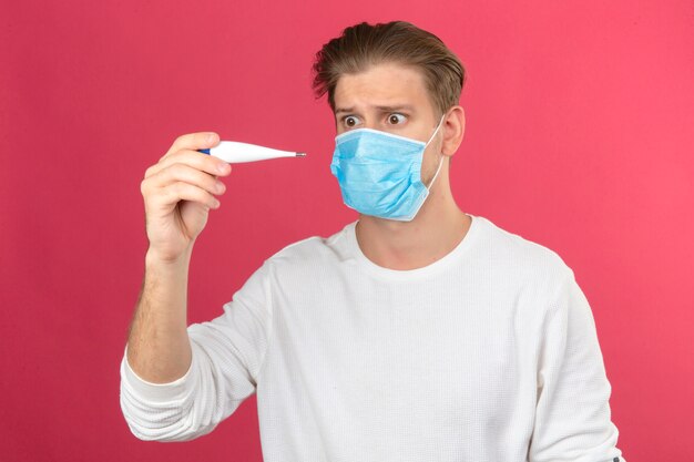 Young man in medical protective mask looking at digital thermometer in panic shocked and surprised over isolated pink background