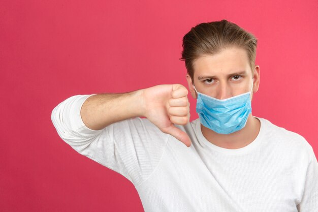 Young man in medical protective mask looking sad making thumbs down showing dislike over isolated pink background