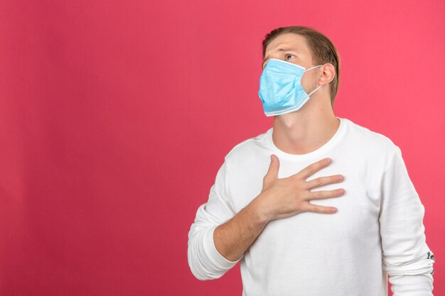 Young man in medical protective mask looking sick and frightened touching his chest over isolated pink background with copy space