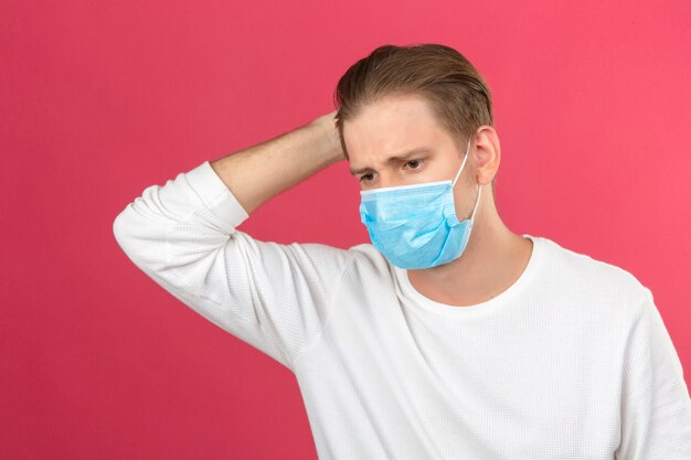 Young man in medical protective mask looking sick touching head standing over isolated pink background