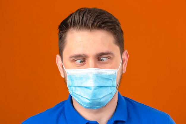 Young man in medical protective mask making grimace crossing his eyes over isolated orange wall