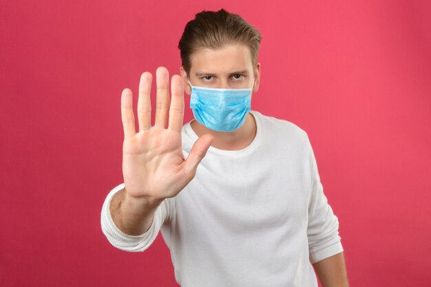 Young man in medical protective mask making stop sign with hand looking at camera with serious face standing over isolated pink background