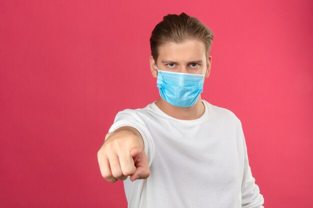 Young man in medical protective mask pointing finger to camera with serious face standing over isolated pink background