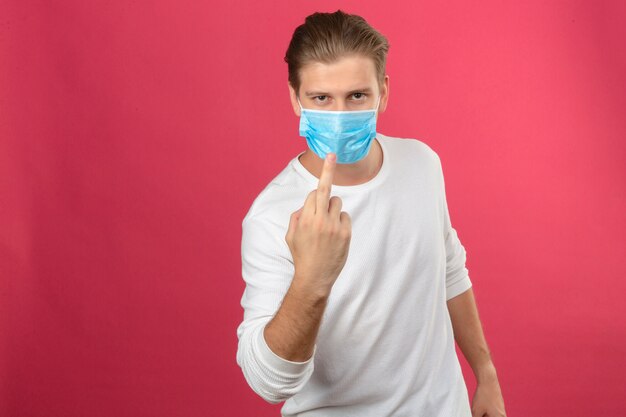 Young man in medical protective mask showing middle finger rude expression over isolated pink background