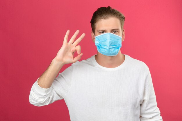 Young man in medical protective mask showing ok sign with fingers and hand looking at camera over isolated pink background