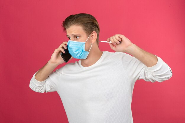 Young man in medical protective mask speaking by smartphone in panic over isolated pink background