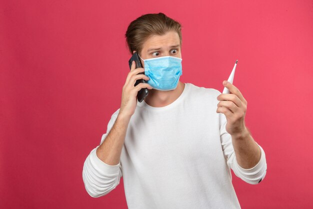 Young man in medical protective mask speaking by smartphone in panic and looking at thermometer over isolated pink background