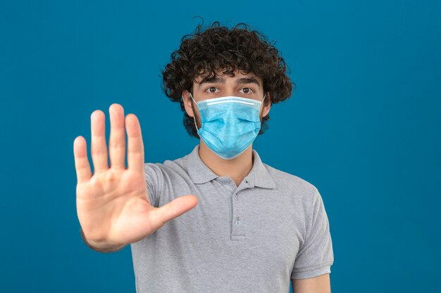 Young man in medical protective mask standing with open hand doing stop sign with serious and confident expression defense gesture over isolated blue background