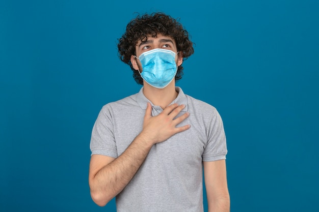 Young man in medical protective mask touching on chest to check lung while breathing over isolated blue background