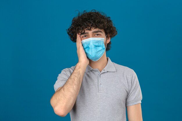 Молодой человек в медицинской защитной маске трогает голову, выглядит больным и имеет головную боль, стоя с несчастным лицом на изолированном синем фоне