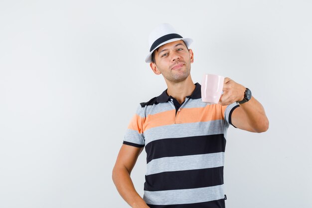 Young man offering cup of tea in t-shirt and hat and looking polite