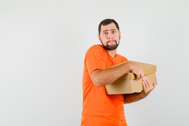 Young man opening cardboard box in orange t-shirt and looking serious , front view.