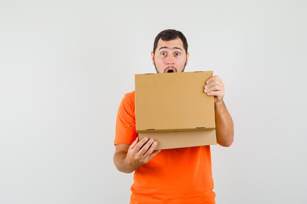 Young man opening cardboard box in orange t-shirt and looking surprised. front view.