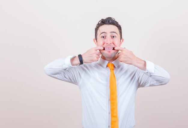 Young man opening mouth with fingers, sticking out tongue in shirt and looking funny